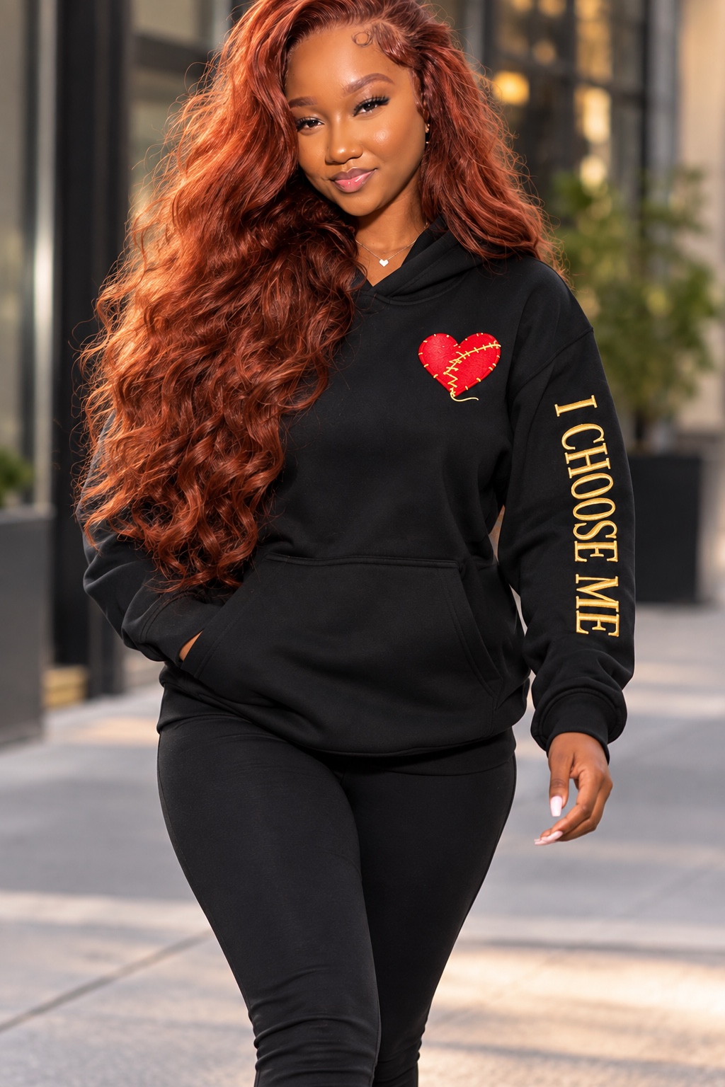 Model wearing the INFIDELITY HURTS I CHOOSE ME black hoodie with mended heart embroidery and gold sleeve lettering